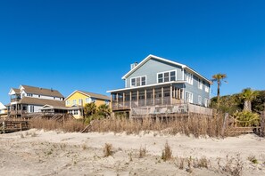 A beachfront property with a large house featuring a balcony, a wooden deck, and a sandy beach in the foreground.