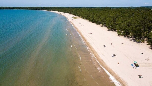 Vue aérienne d’une plage aux eaux turquoise et limpides, bordée d’un rivage sablonneux et d’une forêt dense sur la droite.