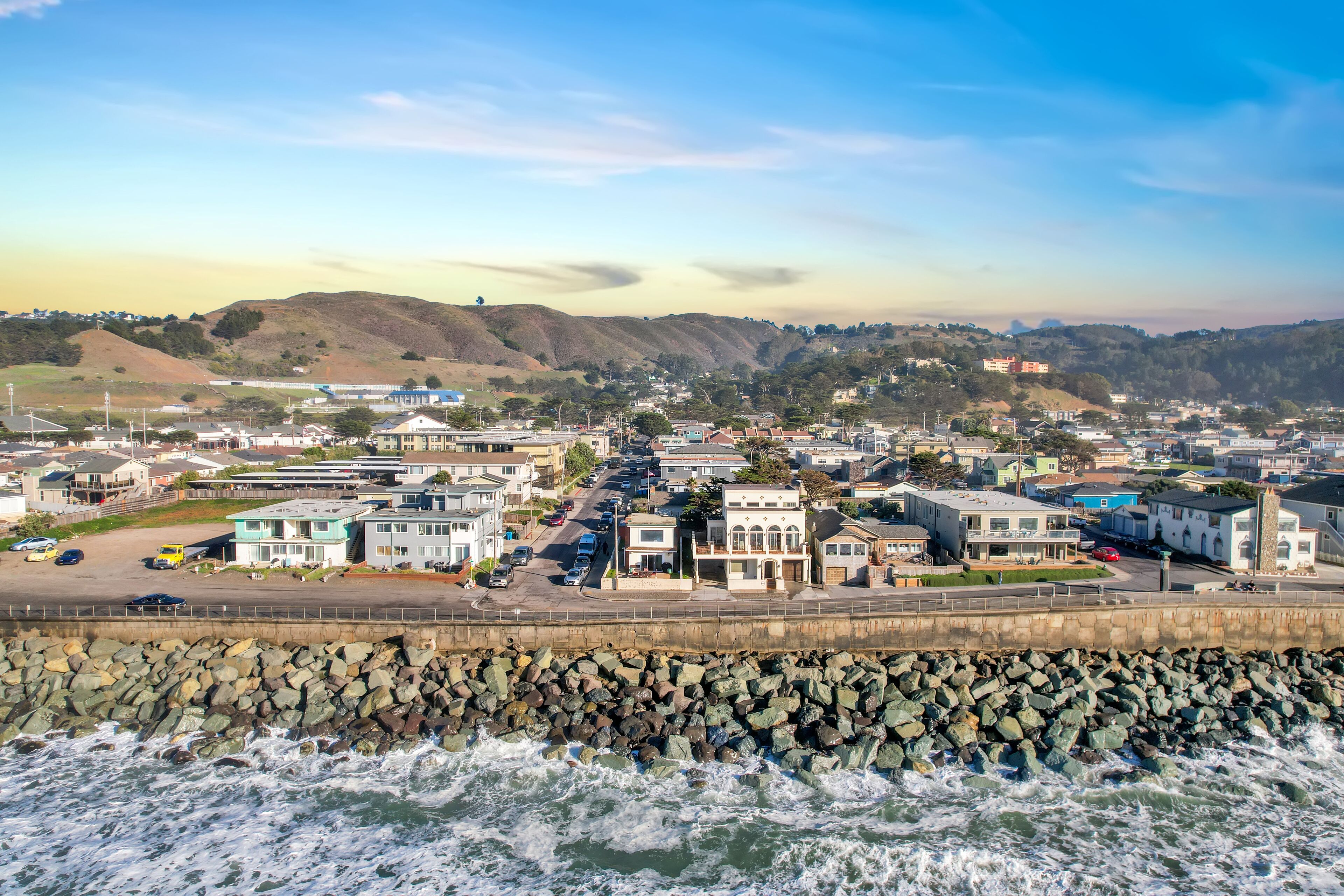 A coastal town with a rocky breakwater, residential buildings, and hills in the background.
