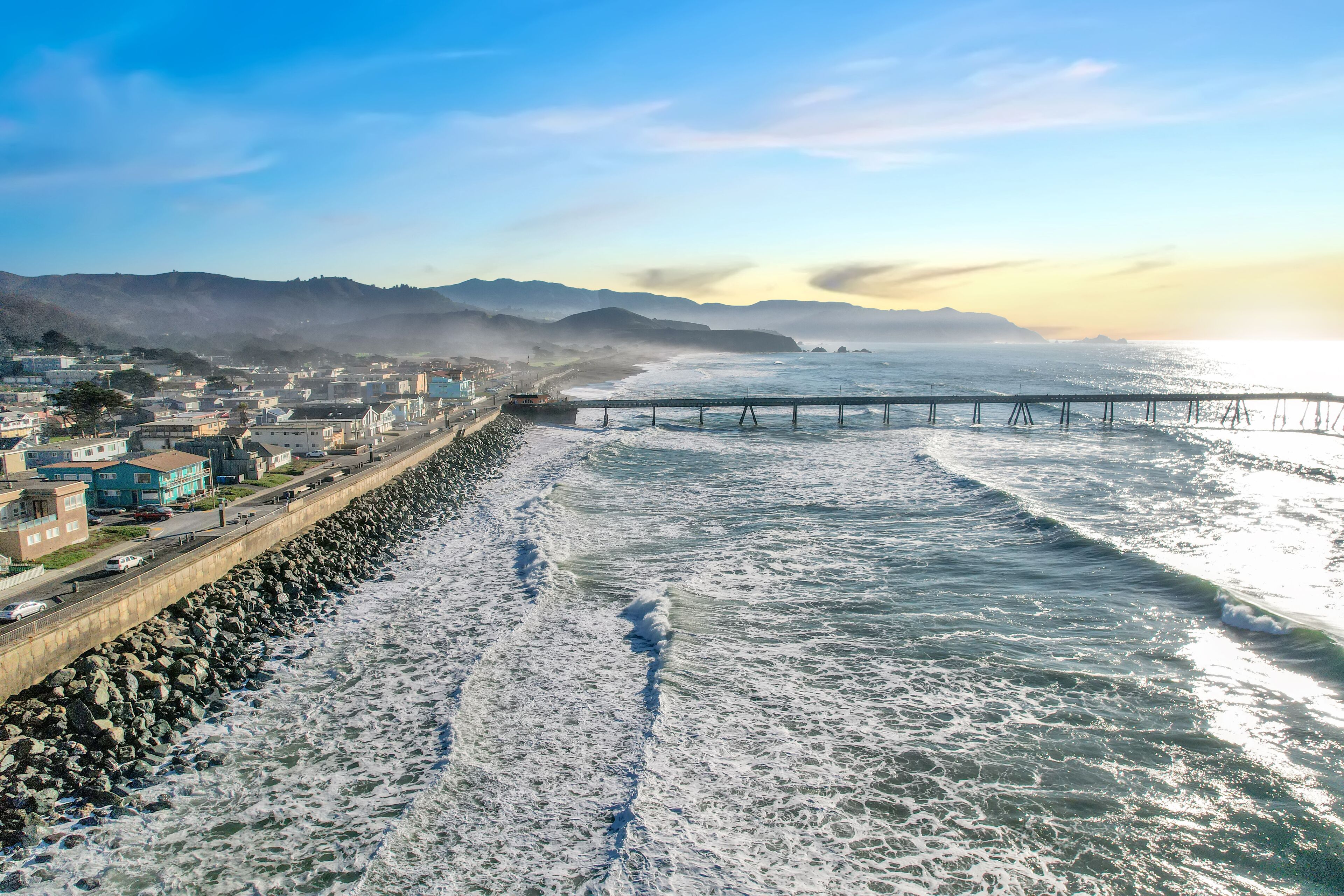 A coastal cityscape with a long pier extending into the ocean, a rocky shoreline, and buildings along the coast.