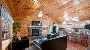A cozy cabin interior with a fireplace, wooden ceiling, black leather sofas, a stone fireplace, and a kitchen area with wooden cabinets and bar stools. - Close ‘n Secluded❤️Mountain Views*PRIVATE RESORT*Hot Tub*Cabin😊A+++ (Sevierville)