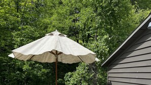 Eine Holzterrasse mit einer weißen Rattan-Couch, einem kleinen Tisch, einem Sonnenschirm und einem hölzernen Geländer.