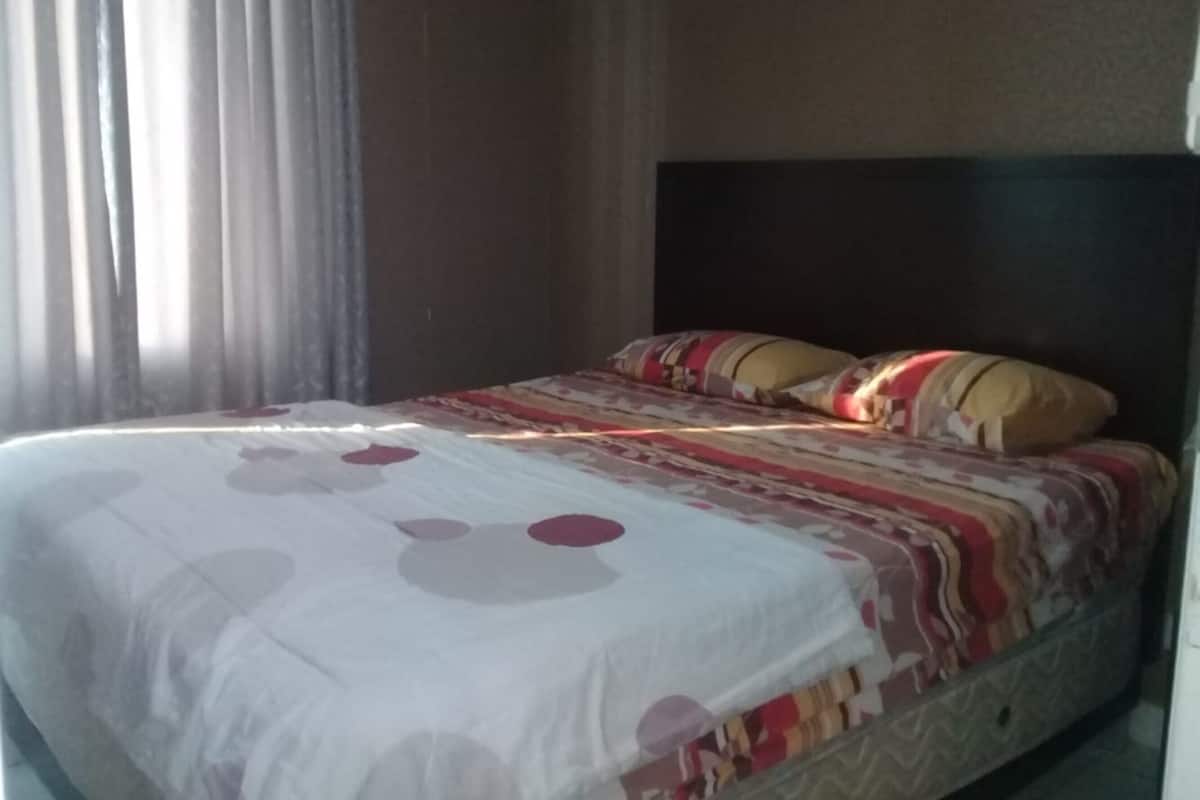 Uma cama bem arrumada com colcha estampada e travesseiros, cabeceira escura e uma janela com cortinas.