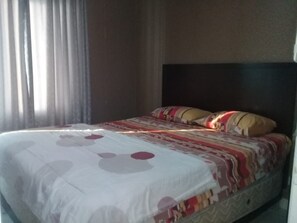 A bed with a patterned bedspread and pillows, a dark headboard, and a window with curtains. - Adaru Property @city Home Apartmen KLP GDG by Zuzu (Jakarta)