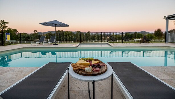 Un espace aménagé au bord de la piscine, avec des chaises longues, une table sur laquelle est disposé un plateau de fromages, et une vue sur le coucher du soleil.