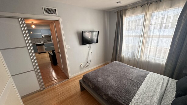 Ein Schlafzimmer mit einem Bett, einem an der Wand montierten Fernseher, einem Fenster mit Vorhängen und einer Tür zu einem Badezimmer.
