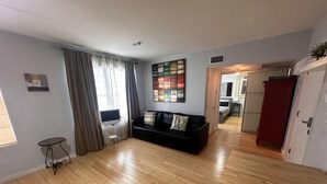 Ein geräumiges Wohnzimmer mit einer schwarzen Couch, Holzboden, einem Fernseher auf einem kleinen Tisch und einem Gemälde an der Wand.