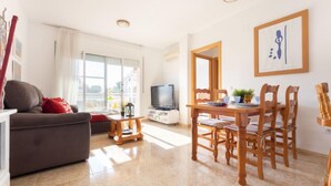 A living room with a sofa, a dining table with chairs, a TV, and a window with a view. - Roquemar by Interhome (L'Ampolla)
