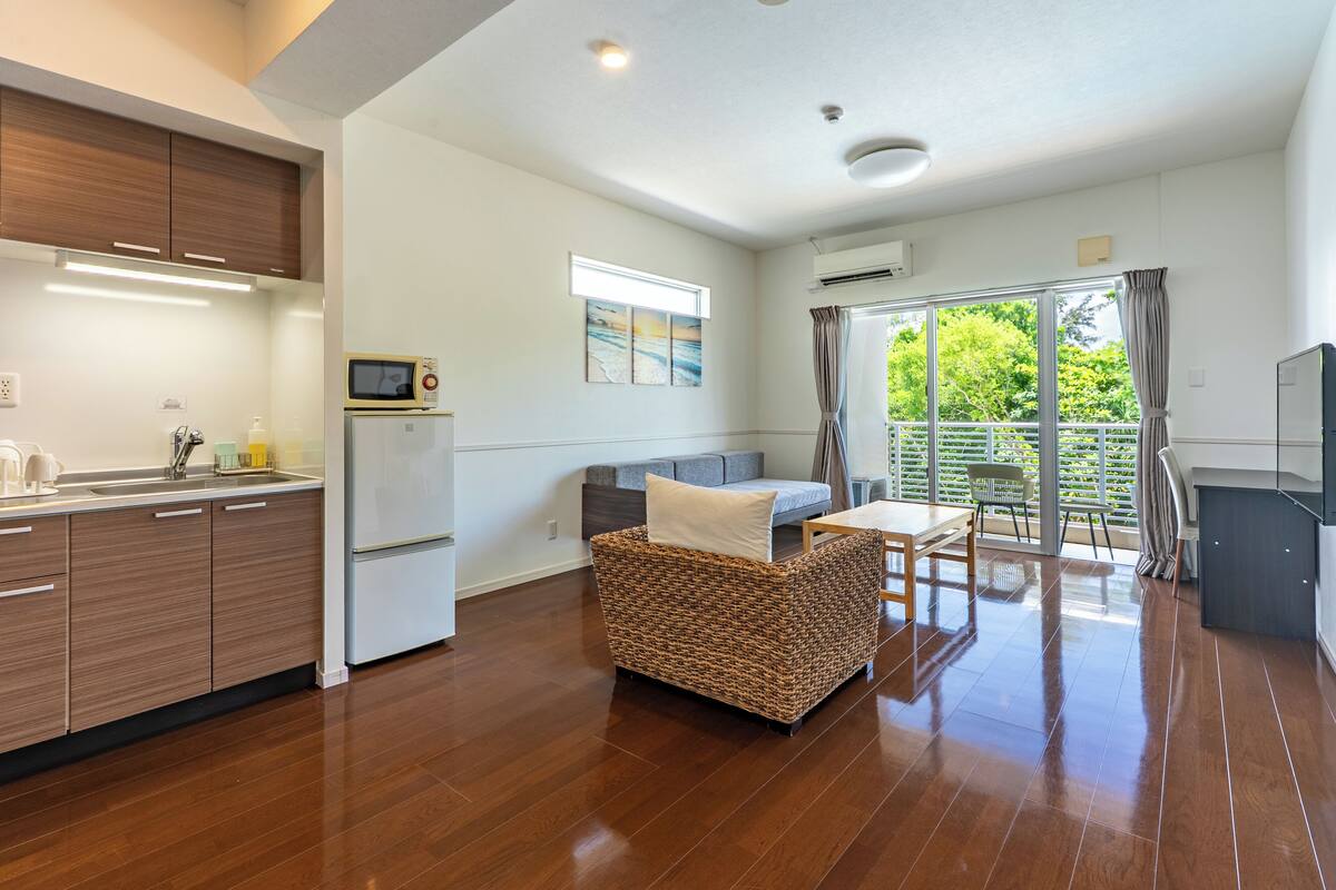 A modern kitchen with wooden cabinets, a refrigerator, and a microwave. There is a wicker sofa with a cushion, a dining table with chairs, and a balcony with a view of greenery.