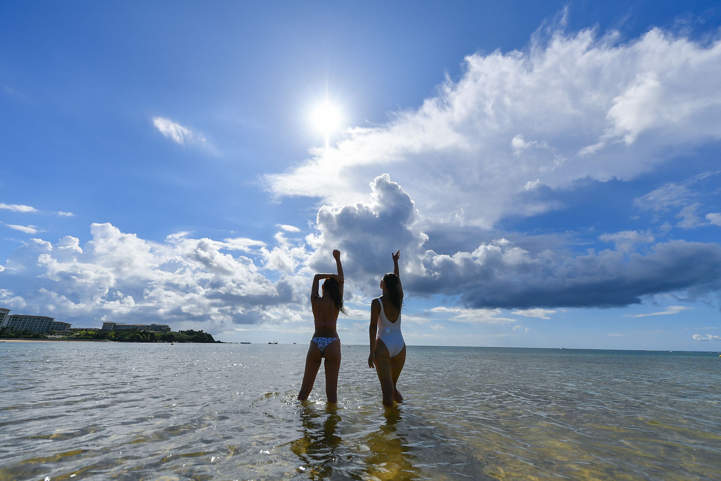 Two people standing in shallow water at a beach with buildings in the background.