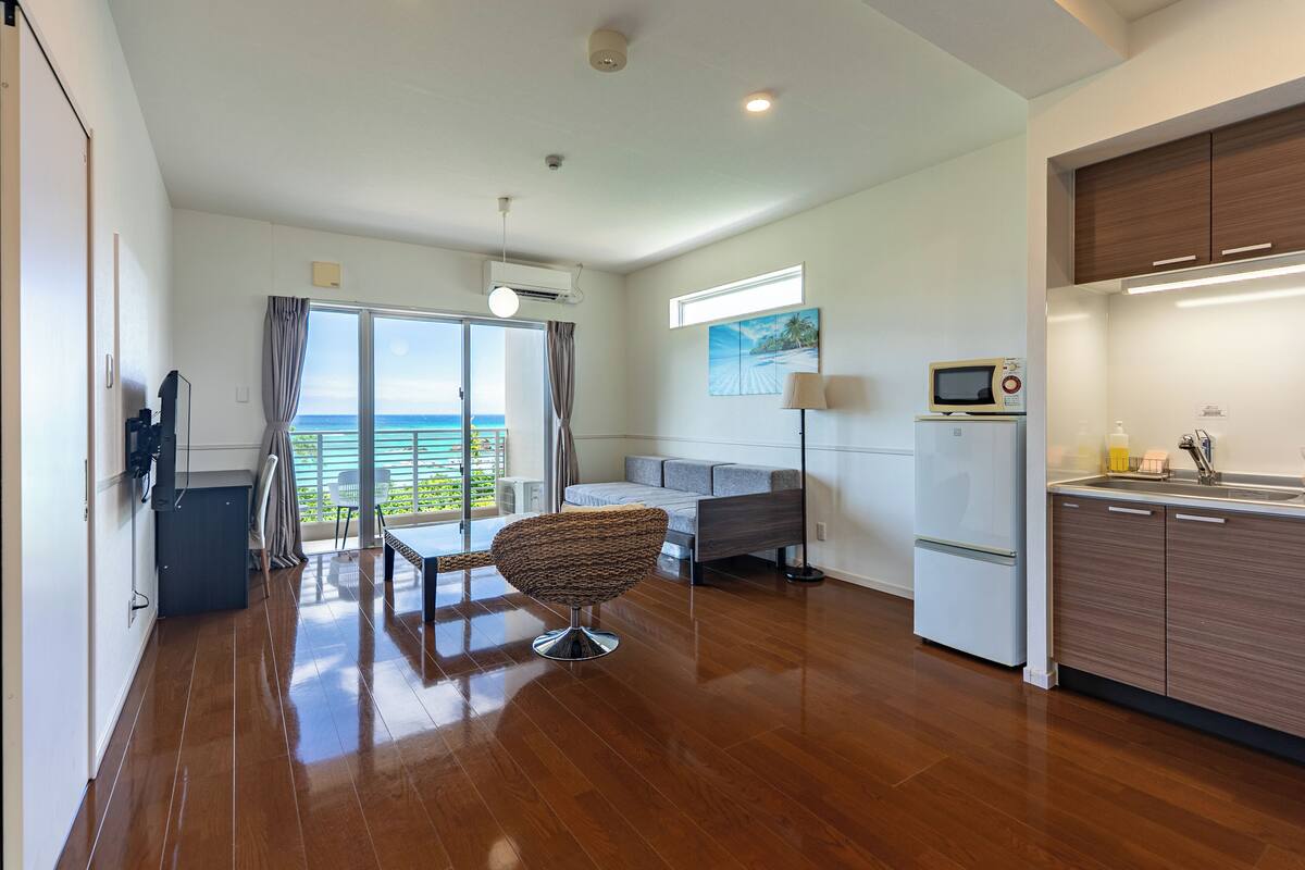 A modern kitchen with a built-in microwave, a refrigerator, and a sink. There is a dining area with a table and chairs, and a large window offering a view of the ocean.