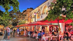 Un espace de restauration en plein air, avec des tables et des chaises placées sous des parasols rouges, entouré de bâtiments dotés d’arcades et d’horloges.