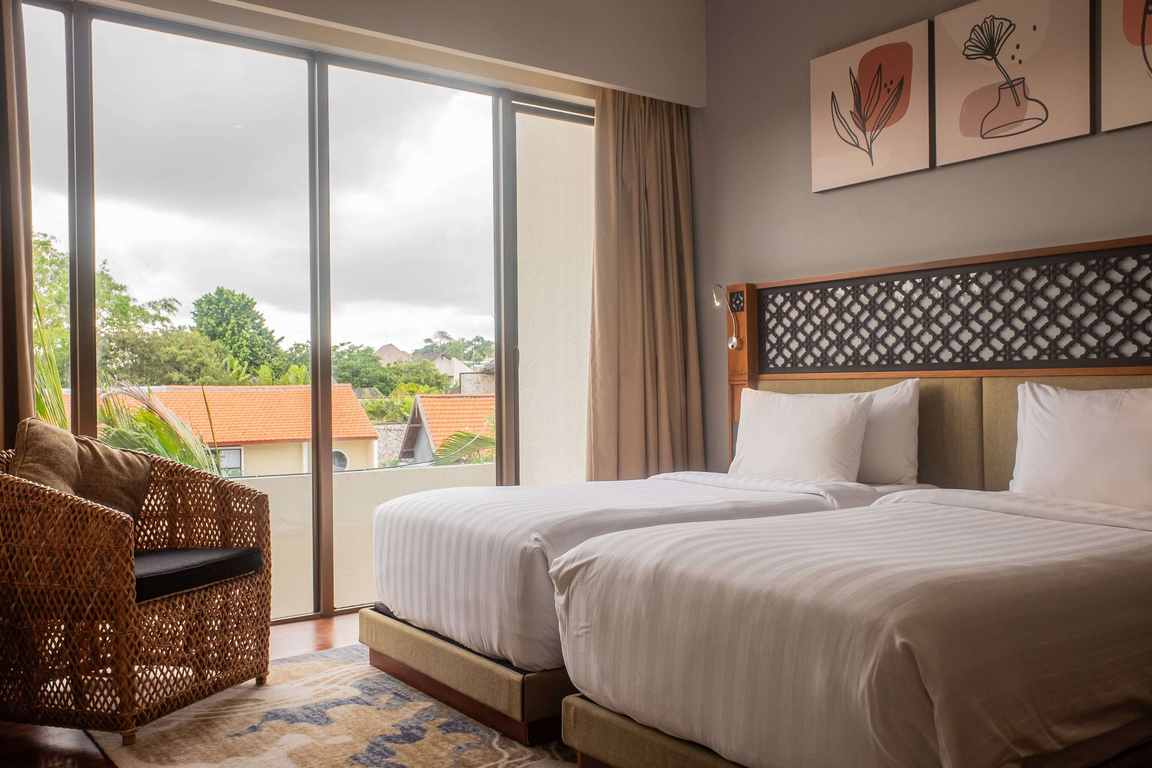 Une chambre d’hôtel avec deux lits, un fauteuil en osier, une grande fenêtre donnant sur un paysage verdoyant et des bâtiments, ainsi que des œuvres d’art accrochées au mur.