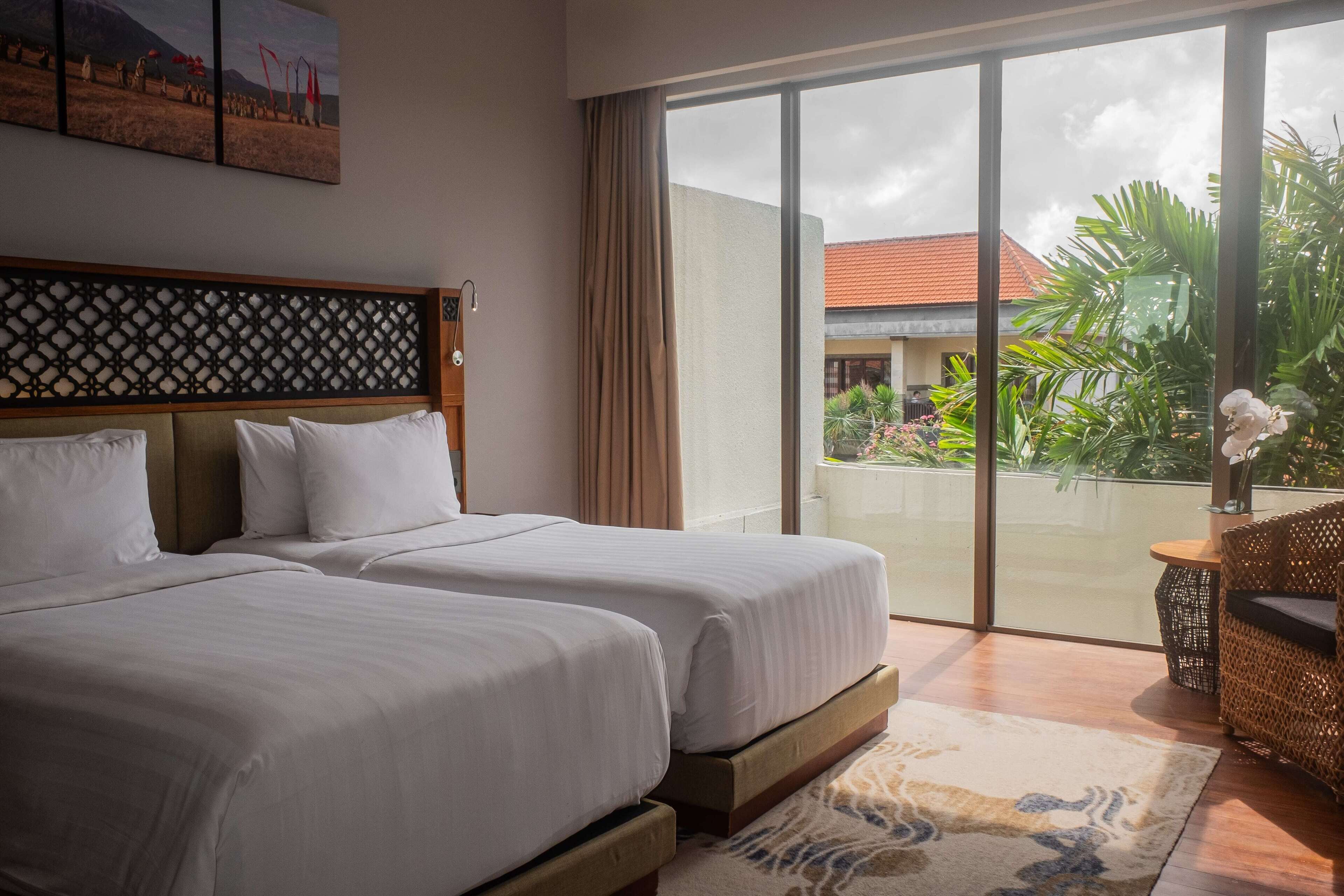 Habitación de hotel con dos camas, un amplio ventanal corredero que da a un balcón con plantas y vistas a un edificio.