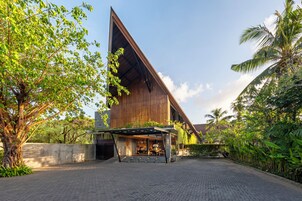 A modern building with a sloped roof, surrounded by lush greenery and a paved entrance area.