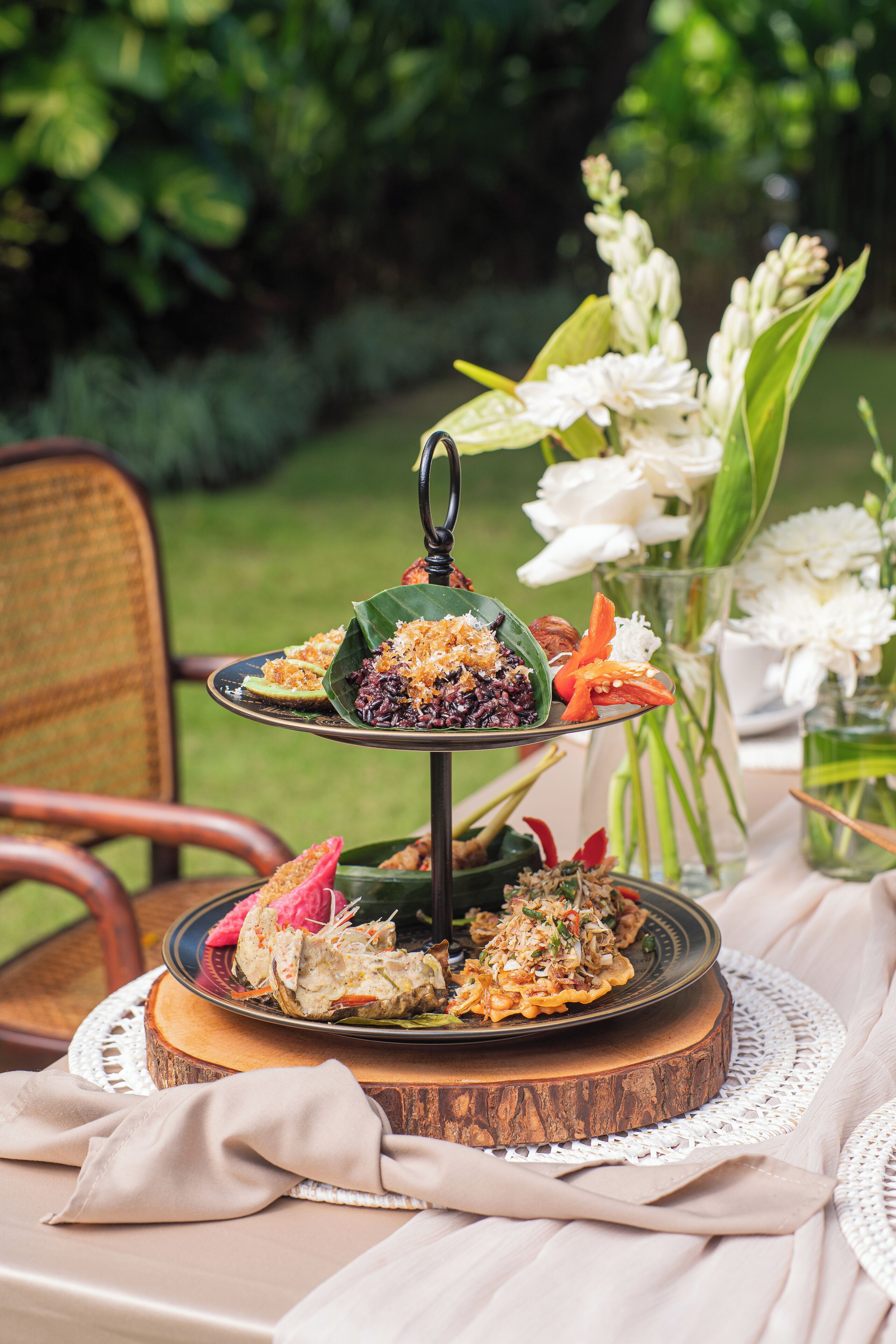 A tiered dessert stand with various sweet dishes, set on a wooden table with a lace doily, surrounded by greenery and white flowers.