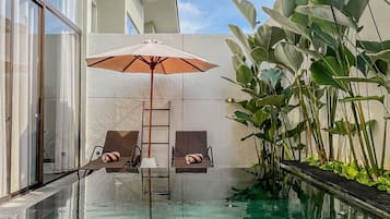 Una piscina infinita con dos sillas reclinables y una sombrilla, rodeada de plantas verdes exuberantes y una fachada de edificio moderno.