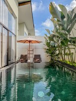 An infinity pool with two lounge chairs and an umbrella, surrounded by large green plants and a modern building facade.