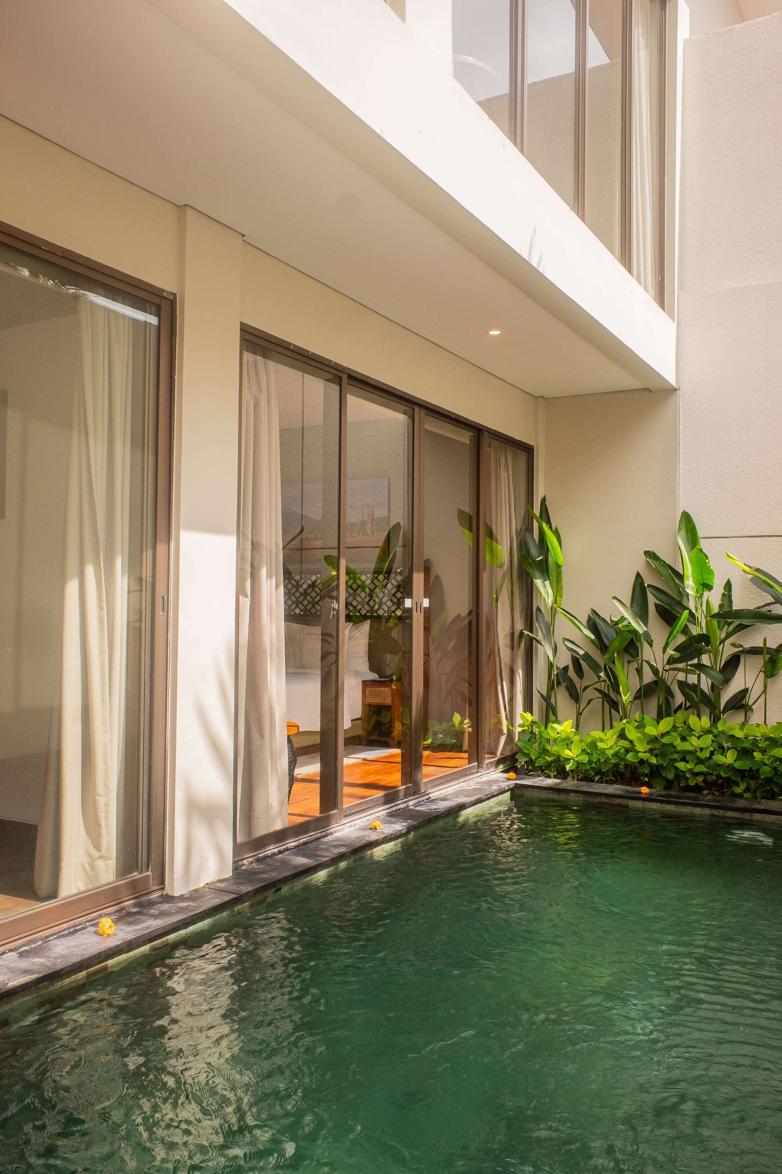 Un área de piscina interior moderna con puertas de vidrio que dan a un salón, rodeada de abundante vegetación.