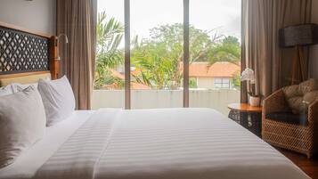 Habitación de hotel con una cama grande, vistas a zonas verdes y edificios, una silla de mimbre y una mesita con una planta.
