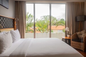 Habitación de hotel con una cama grande, vistas a zonas verdes y edificios, una silla de mimbre y una mesita con una planta.