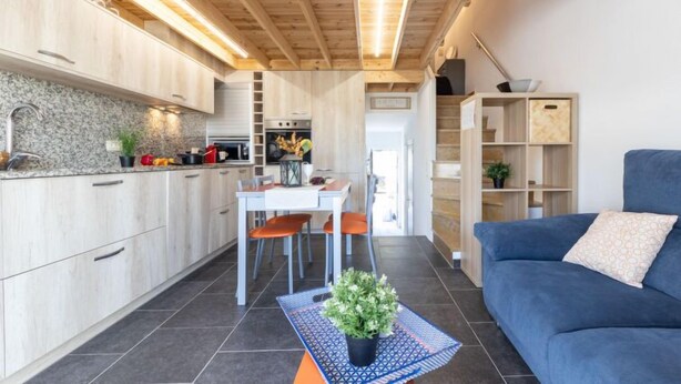 Une cuisine moderne avec des meubles en bois, un coin repas avec une table et des chaises, et un canapé bleu dans l’espace salon.