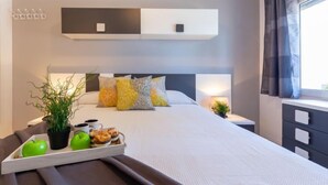 Une chambre d’hôtel moderne avec un lit soigneusement fait, une table de chevet avec une lampe, un plateau avec des pommes et des pâtisseries, et une plante en pot.