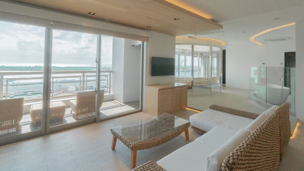 A modern living room with a large glass door leading to a balcony with a view of the sea. There is a sofa, a coffee table, and a flat-screen TV mounted on the wall. - THIRD Ishigaki (Ishigaki)