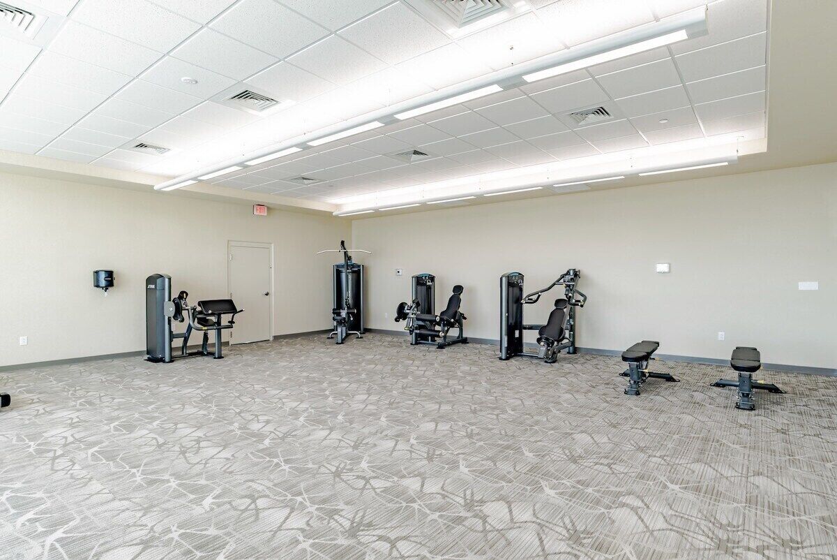 Ein geräumiger Fitnessraum mit verschiedenen Trainingsgeräten, einem gemusterten Teppich und einer Decke mit Neonbeleuchtung.