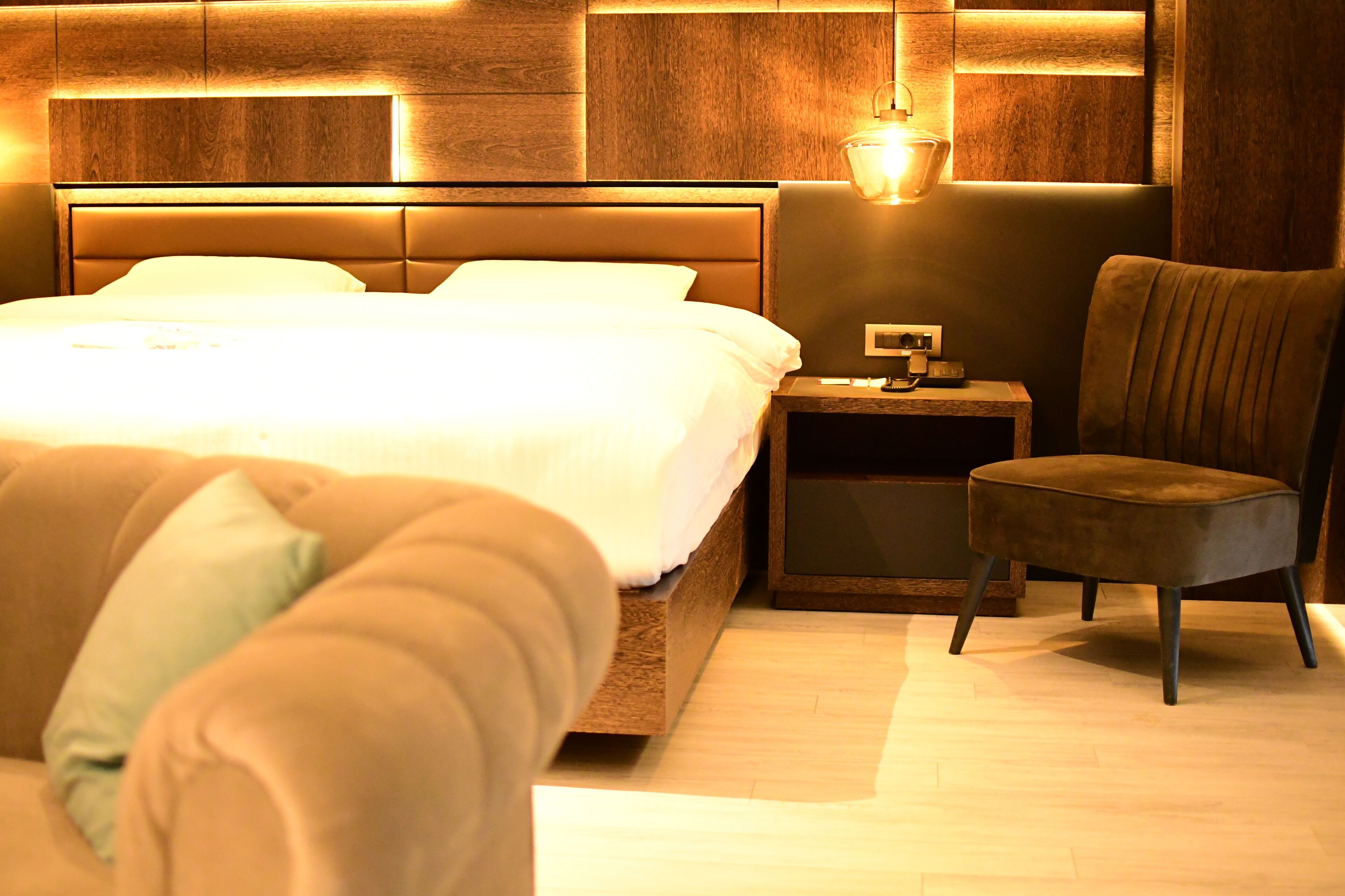 A modern hotel room with a large bed, a nightstand, a chair, and a wall-mounted lamp.