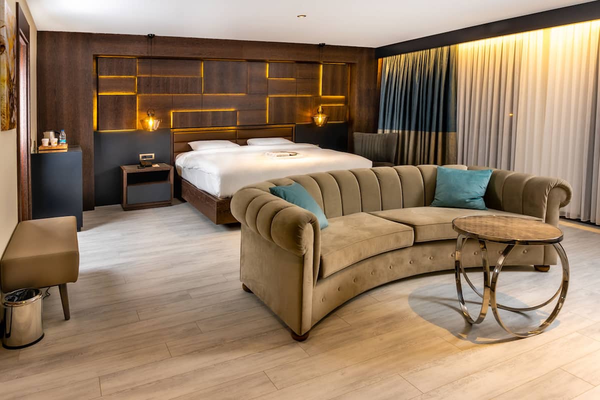 Una habitación de hotel moderna con un sofá curvo, una mesita redonda, una cama con cabecera con paneles iluminados, una mesita de noche con lámpara y un amplio ventanal con cortinas.