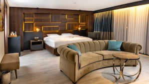 A modern hotel room with a curved sofa, a round side table, a bed with a headboard featuring illuminated panels, a bedside table with a lamp, and a large window with curtains. - Anemon Kent Aydın Otel (Aydin)
