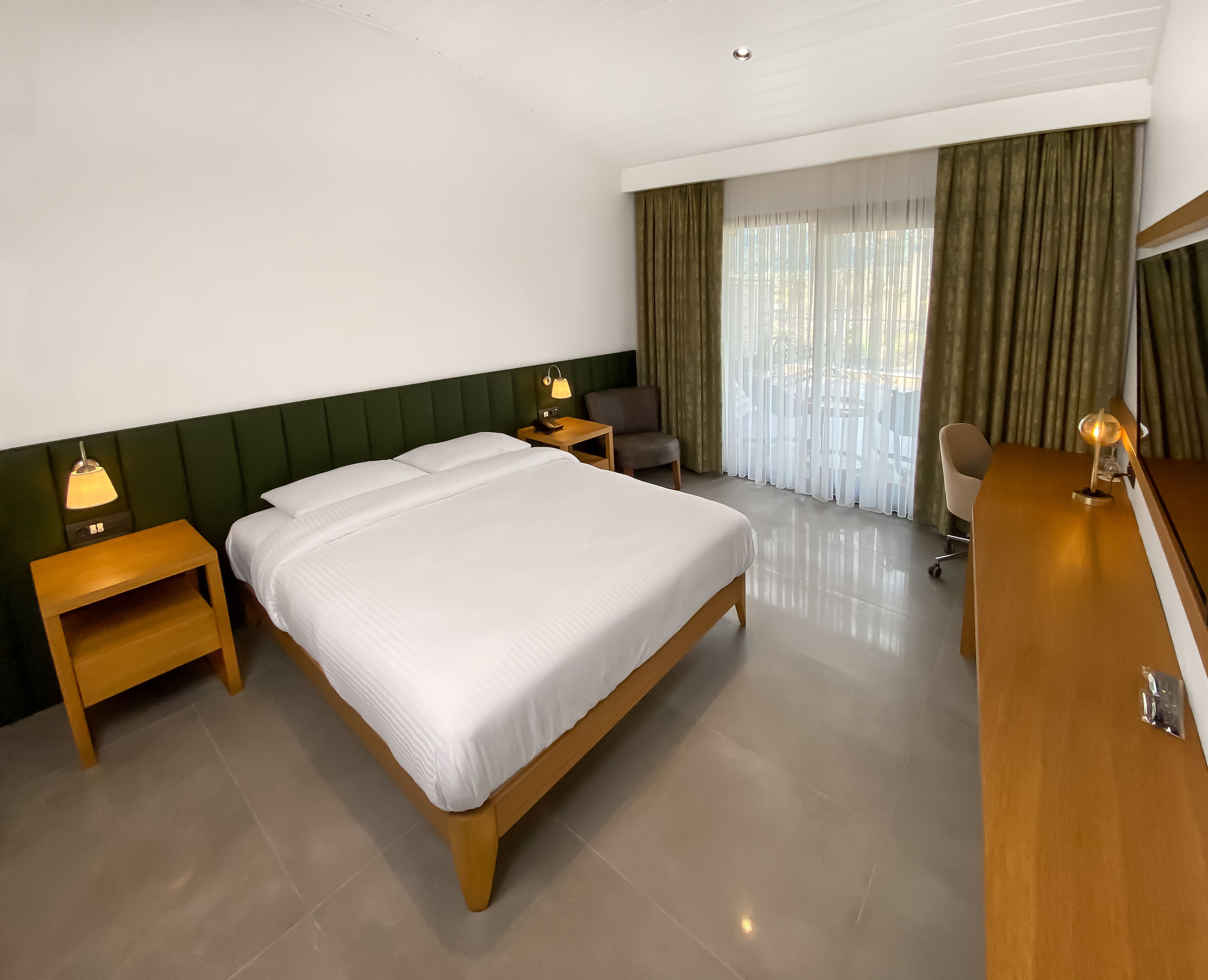 Habitación de hotel con una cama grande, dos mesitas de noche con lámparas, un escritorio con una silla y un amplio ventanal con cortinas.
