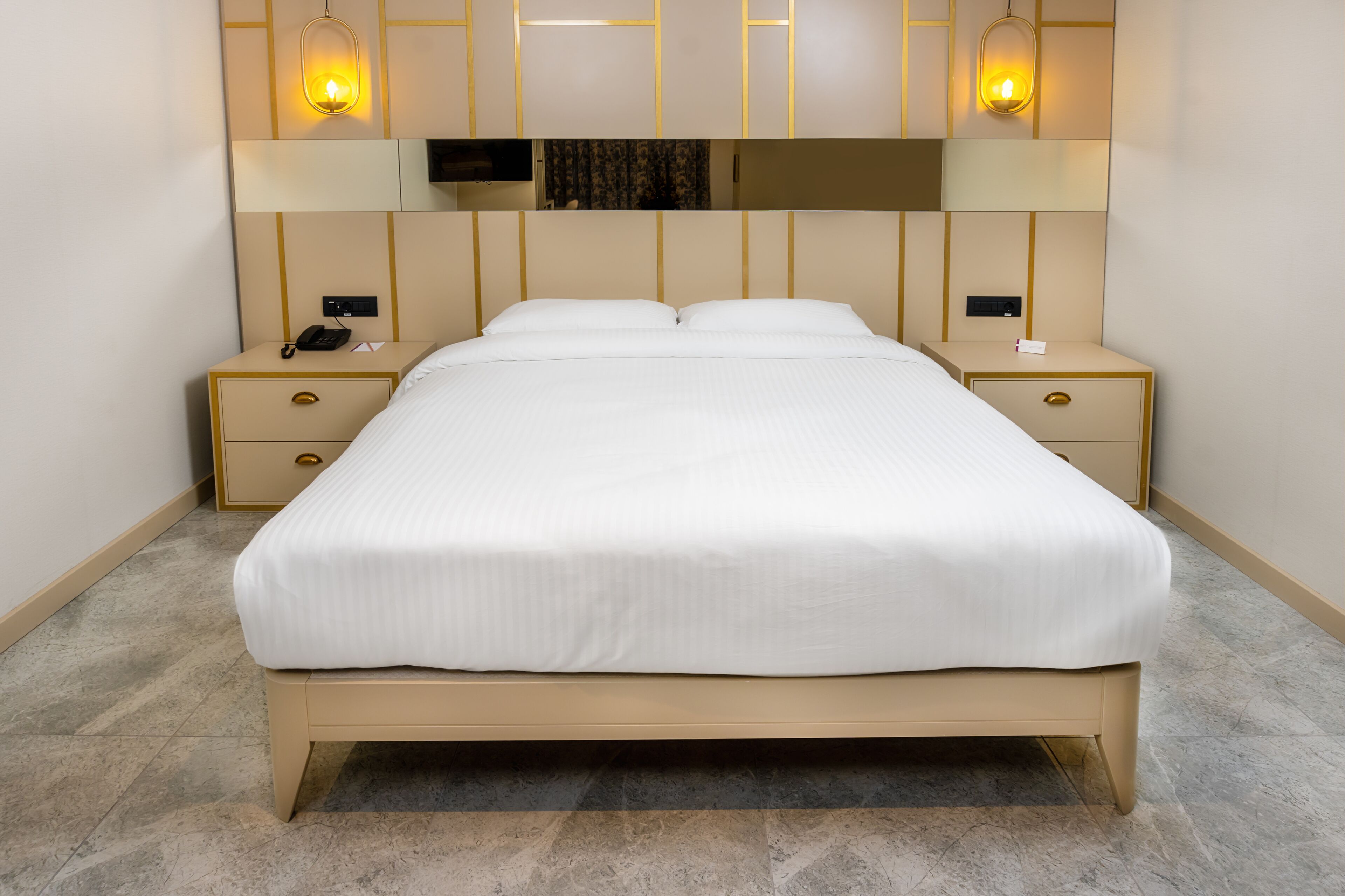 Cama bem arrumada em quarto de hotel com design moderno, incluindo cabeceira com iluminação integrada e pequena mesa de cabeceira com telefone e abajur.