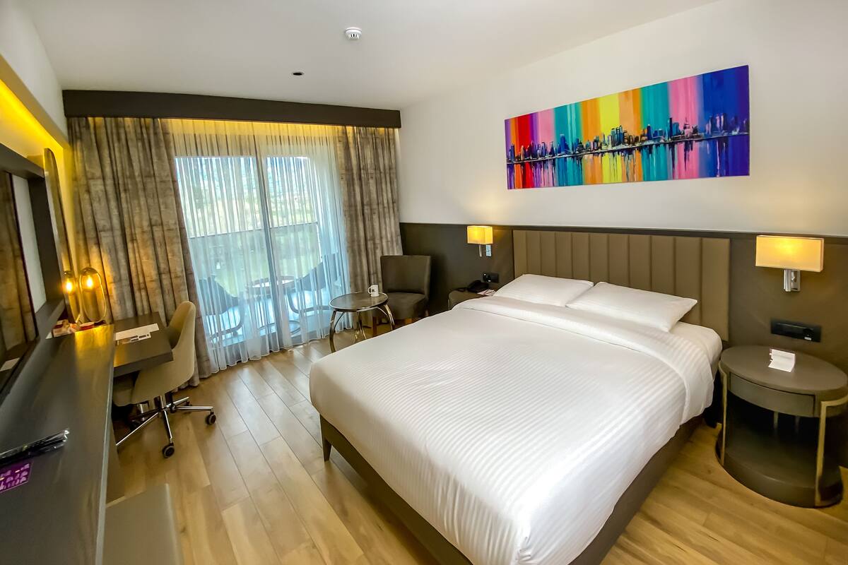 Una habitación de hotel con una cama grande, un escritorio con una silla, una mesita pequeña y otra silla. Hay una ventana con cortinas y un cuadro en la pared.