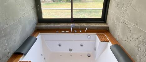 Une salle de bain moderne avec une grande baignoire, des tapis de bain en bois et une vue sur un paysage verdoyant par une grande fenêtre.
