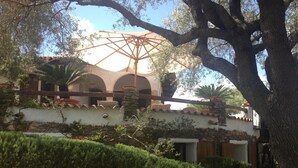 Een gebouw met een witte gevel, een stenen basis en bogen. Op de voorgrond staat een grote boom en er is een pergola.