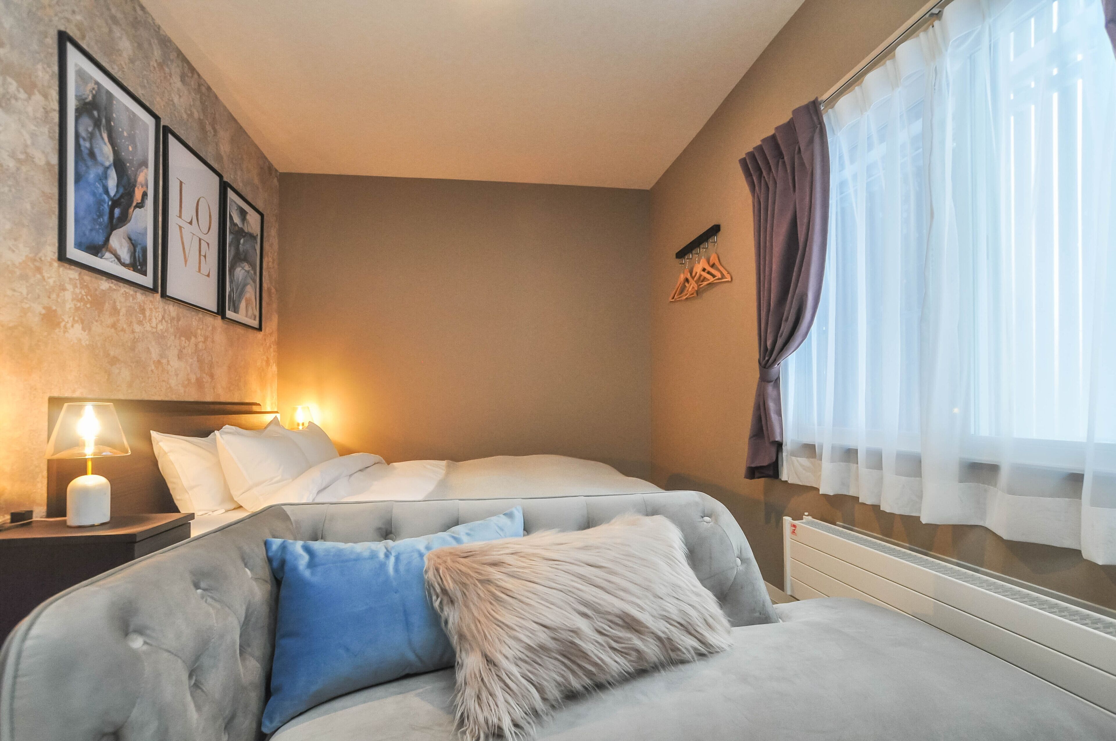 Une chambre avec un lit, une table de chevet avec une lampe, un canapé avec un coussin moelleux et une fenêtre avec des rideaux légers.