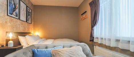 Une chambre avec un lit, une table de chevet avec une lampe, un canapé avec un coussin moelleux et une fenêtre avec des rideaux légers.