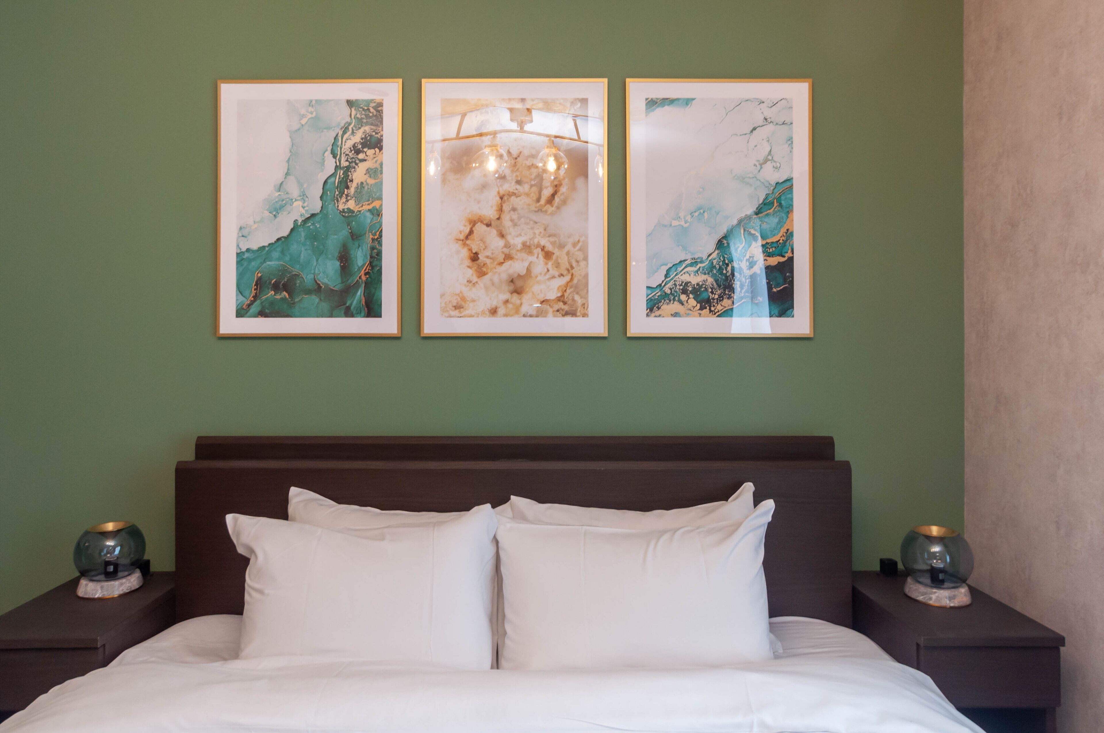 A neatly made bed with white linens and two pillows, flanked by nightstands with lamps. Above the bed, there are three framed abstract paintings on the wall.