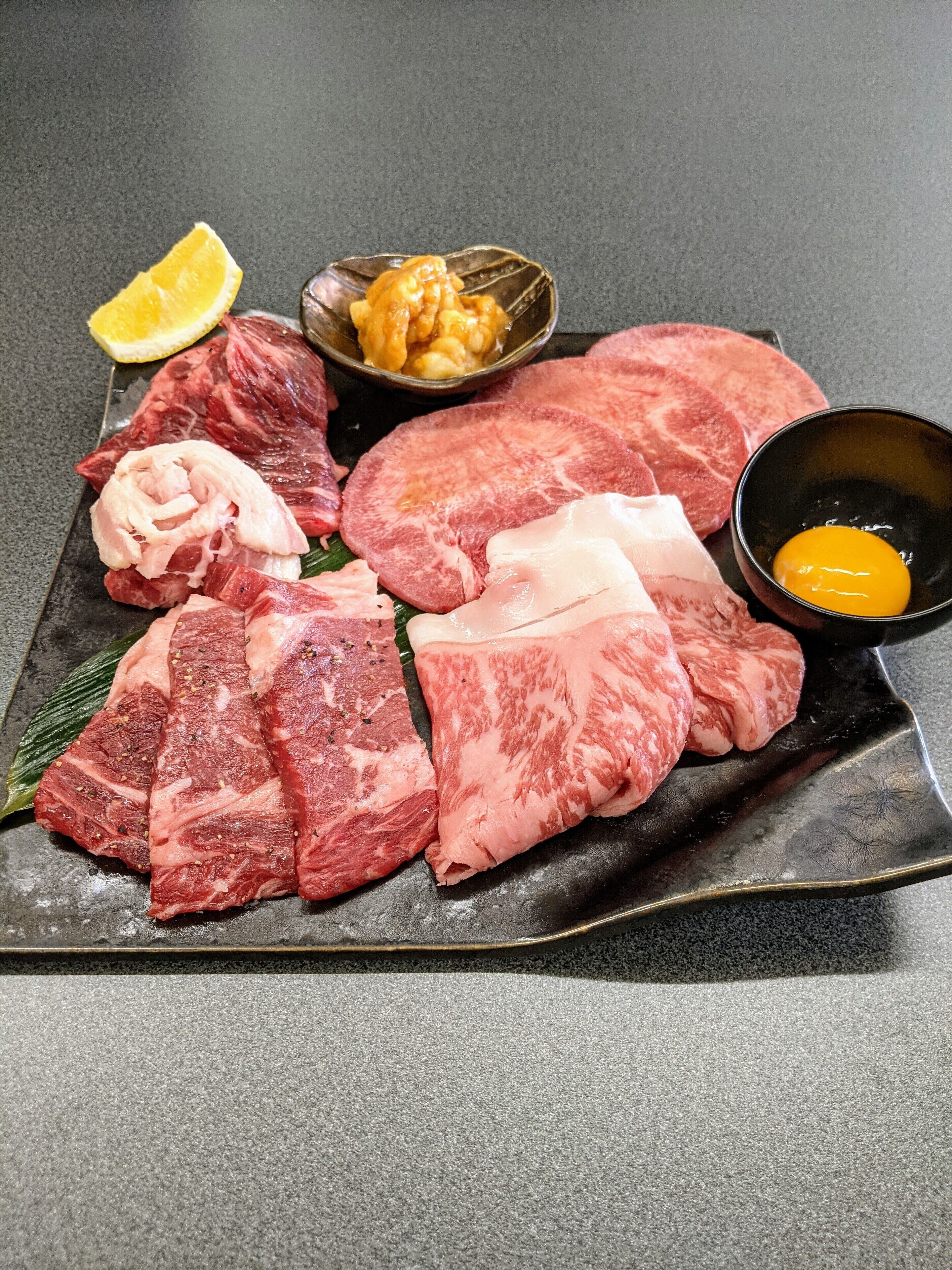 A variety of raw meats on a black plate, including slices of beef, pork, and chicken, with a lemon wedge and a bowl of sauce.