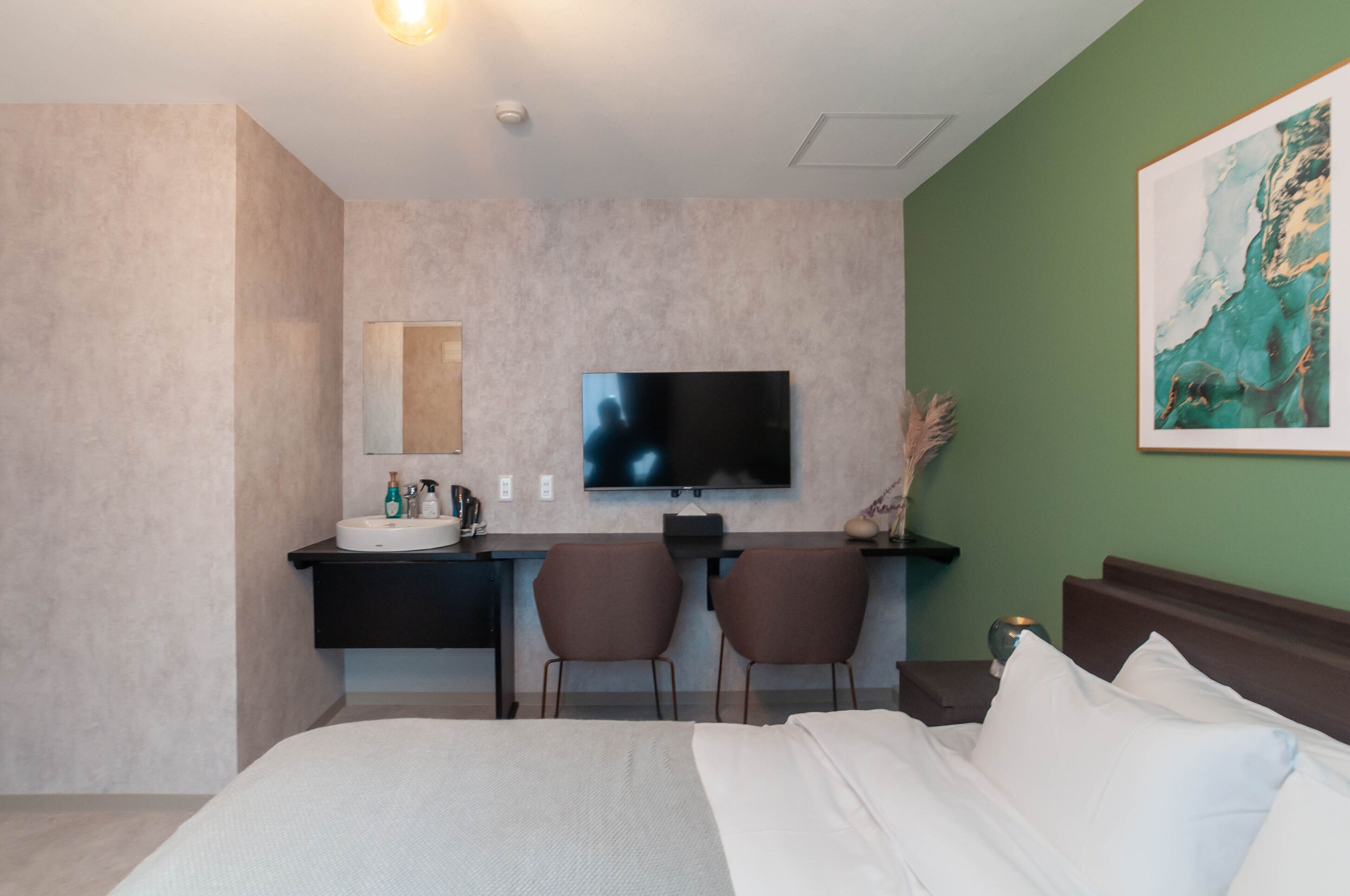 Habitación de hotel con cama, escritorio con lavamanos, dos sillas, televisión de pantalla plana y un cuadro en la pared.