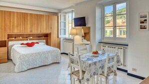 Un dormitorio con cama, mesita de noche, lámpara y vistas a edificios a través de una ventana.