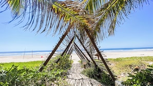 A beach pathway with palm fronds overhead, leading to a sandy beach with ocean in the background.