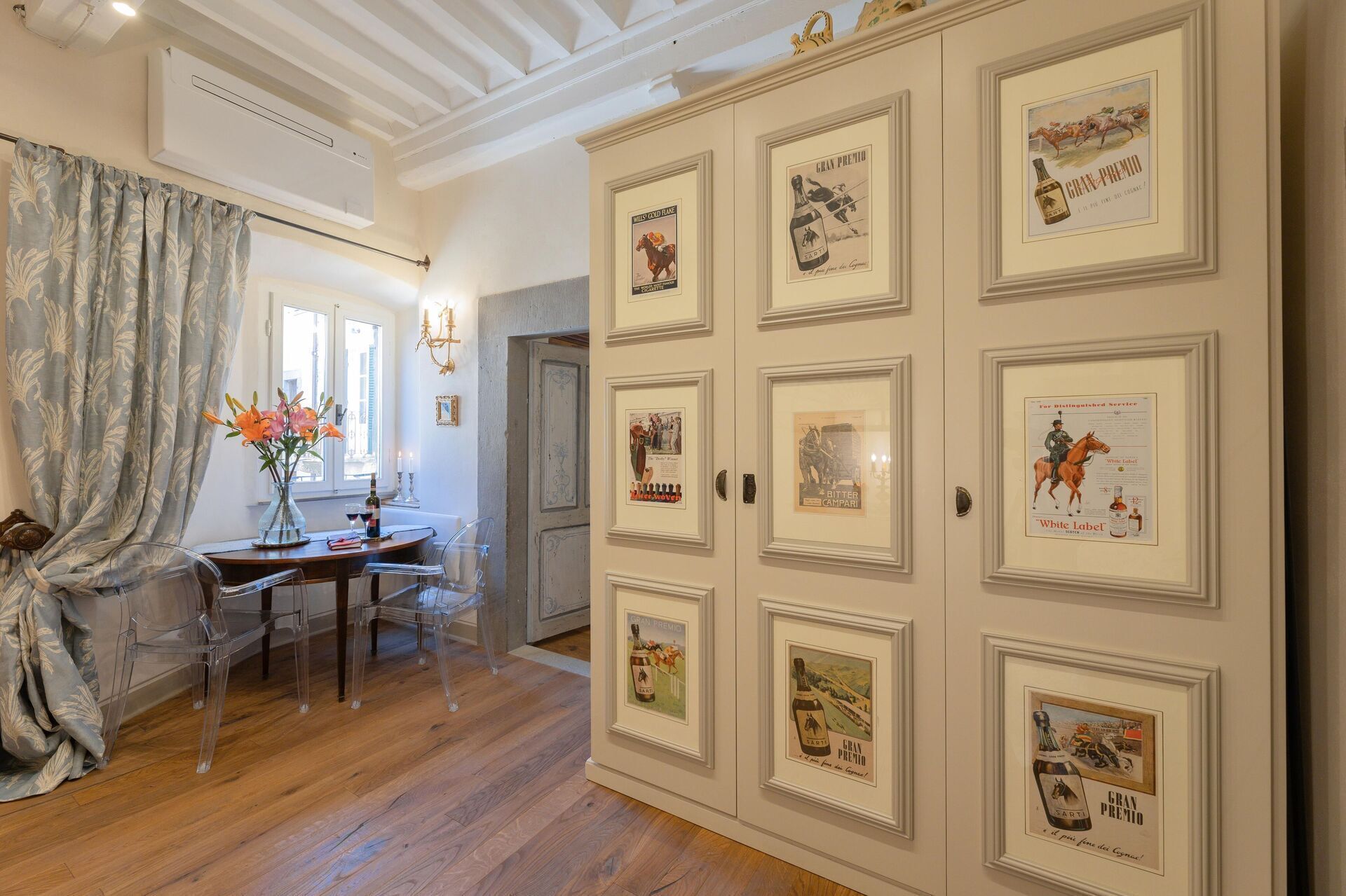 A room with a wooden floor, a dining table with chairs, a vase with flowers, and a wall with framed vintage advertisements.