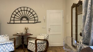 A room with a wooden floor, a round table with a candelabra, two patterned armchairs, a clear glass chair, a white door, and a decorative wall piece. - Cosy apartment for 2 people with WIFI, A/C and TV (Cortona)