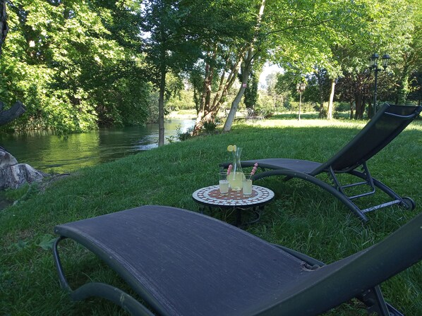 A serene outdoor seating area with two lounge chairs and a small table, set against a backdrop of lush greenery and a calm water body.