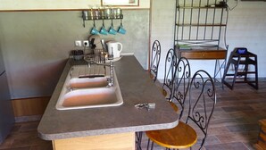 A kitchen with a double sink, a kettle, and a coffee maker. There is a metal rack with cups and a shelf with books and a laptop.