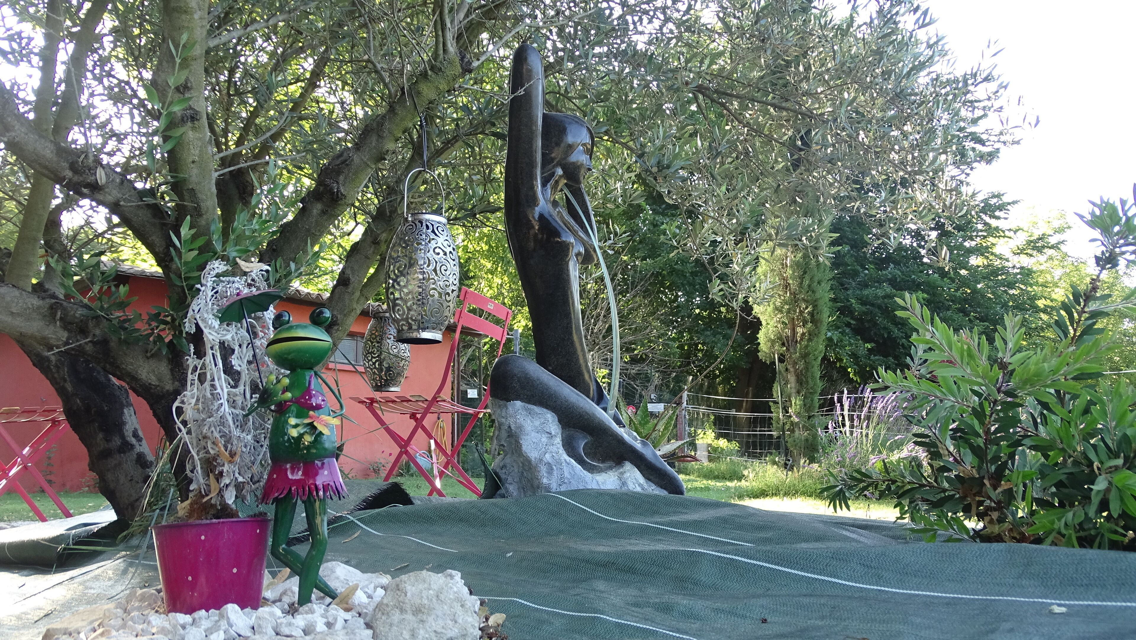 Un jardín de esculturas con diversas instalaciones artísticas, incluyendo una figura de rana y una figura reclinada, rodeado de árboles y una cerca roja.