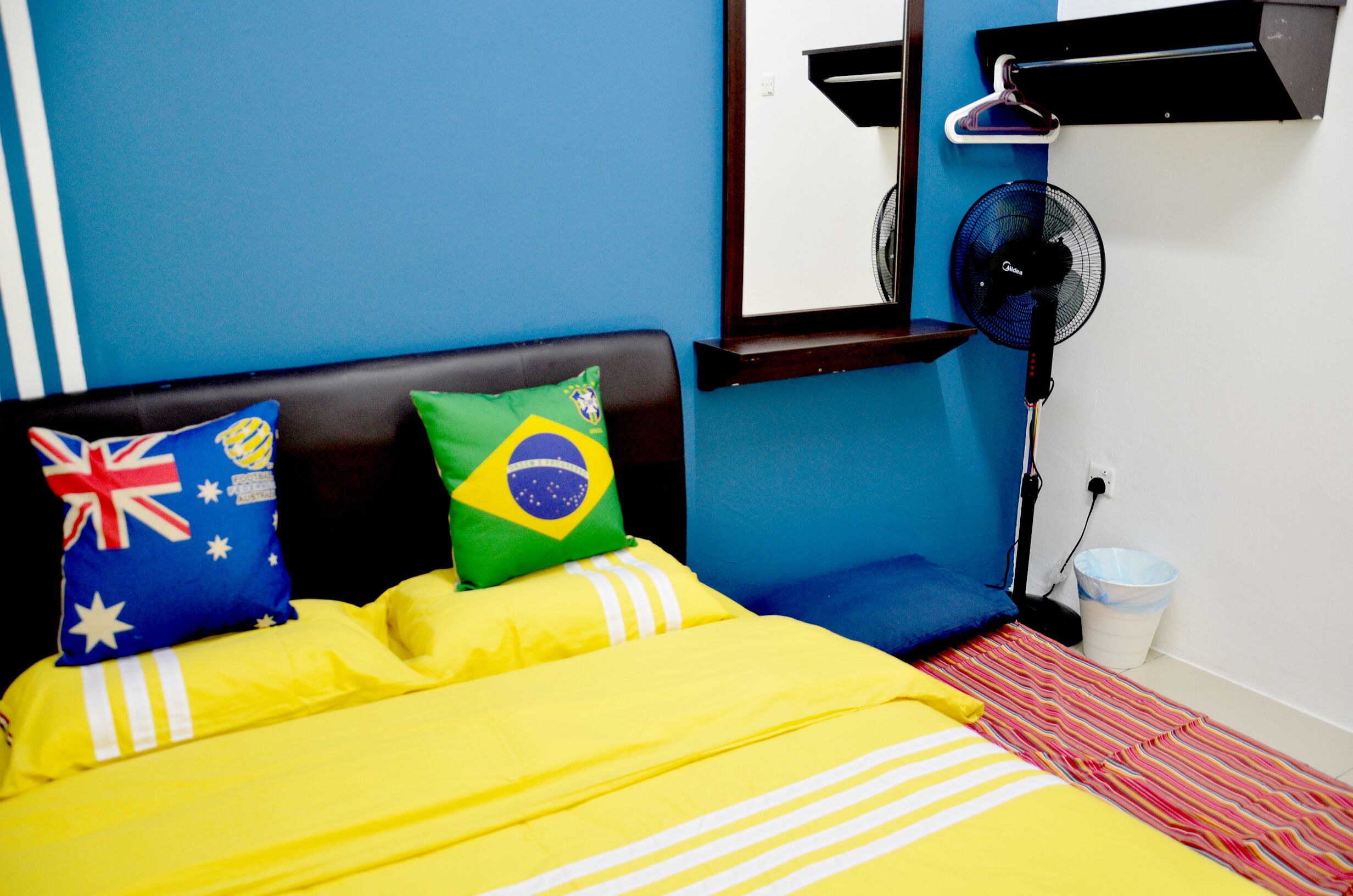 A bed with a yellow bedspread, pillows with flags, a standing fan, and a white trash bin.